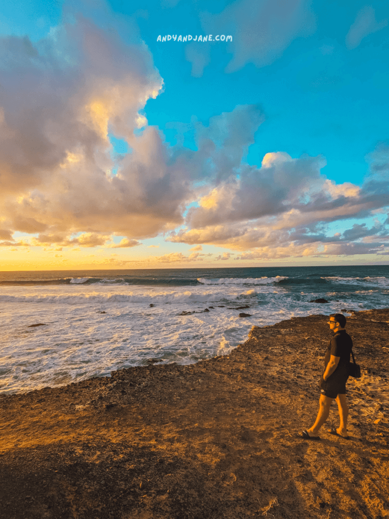 A silhouette of a person stands on rocky terrain by the ocean in La Pared, Fuerteventura, gazing at the vibrant sunset and dramatic clouds above.