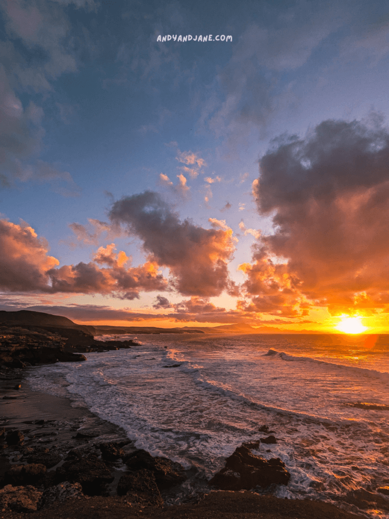 A stunning sunset in La Pared, Fuerteventura, looking over rocky coastline, with waves crashing against the shore and vibrant orange and pink clouds filling the sky.