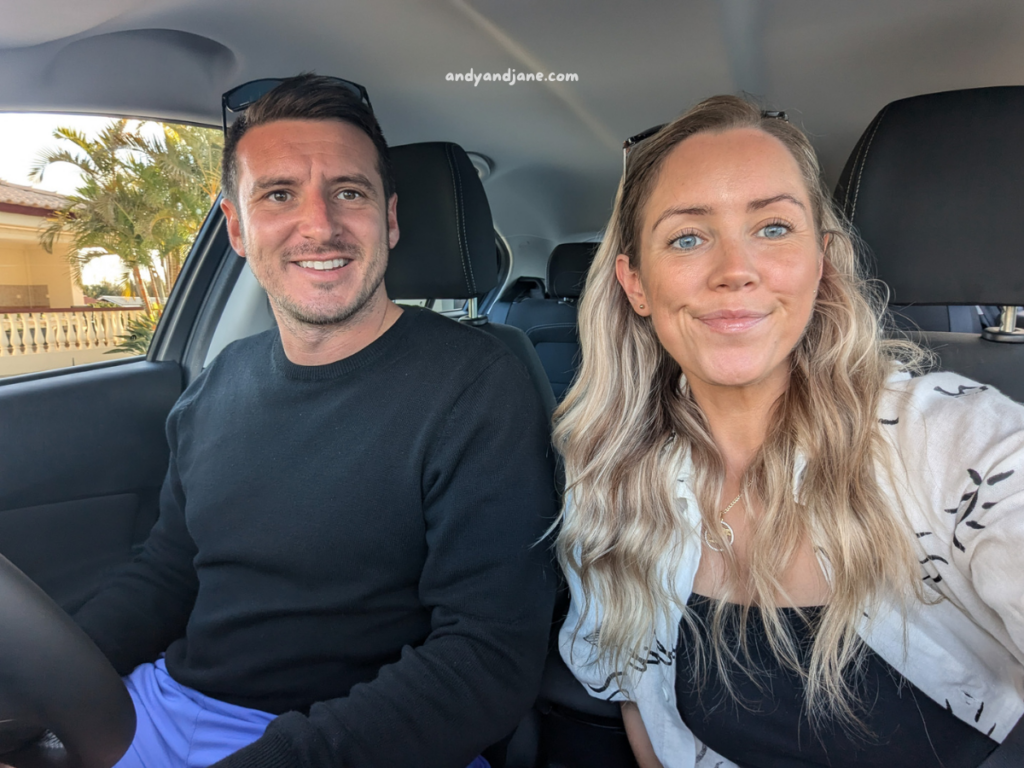 A man and woman sit inside a car, smiling. Sunlight filters through the window, highlighting palm trees outside.