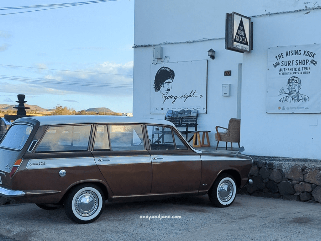 A classic brown station wagon parked outside a surf shop with modern signage, showcasing a coastal vibe against scenic surroundings in Lajares, Fuerteventura