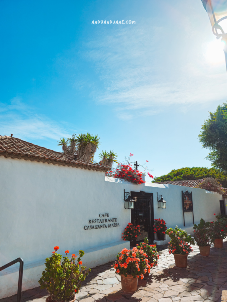 White building with a tiled roof under the clear blue Betancuria sky. Entrance displays "Cafe Restaurante Casa Santa Maria." Red flowers in pots line the stone pathway, basking in the bright sunlight.