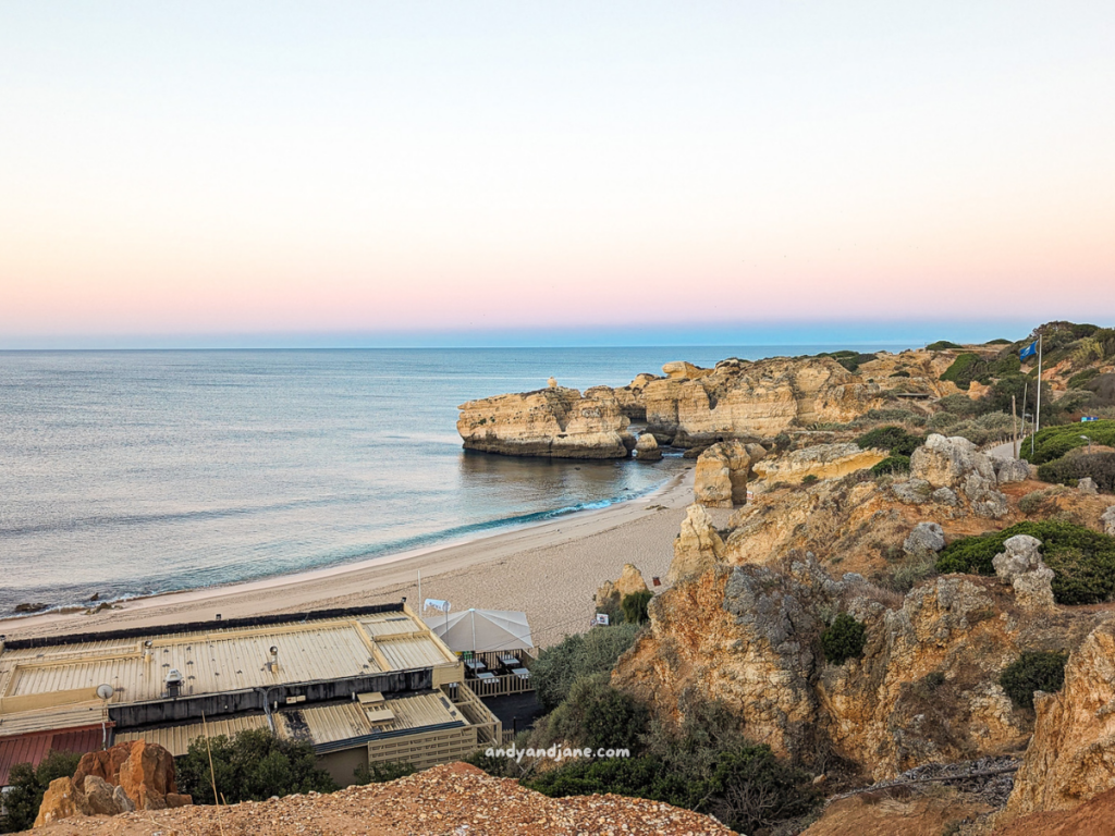 Serene coastal view at sunset in the Algarve, featuring sandy beach, rocky formations, and a beachside structure surrounded by greenery.