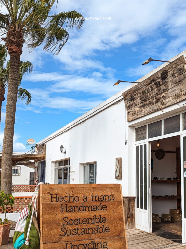 A wooden sign outside a shop reads “Handmade, Sustainable, Upcycling” beside palm trees and blue skies in Lajares town.