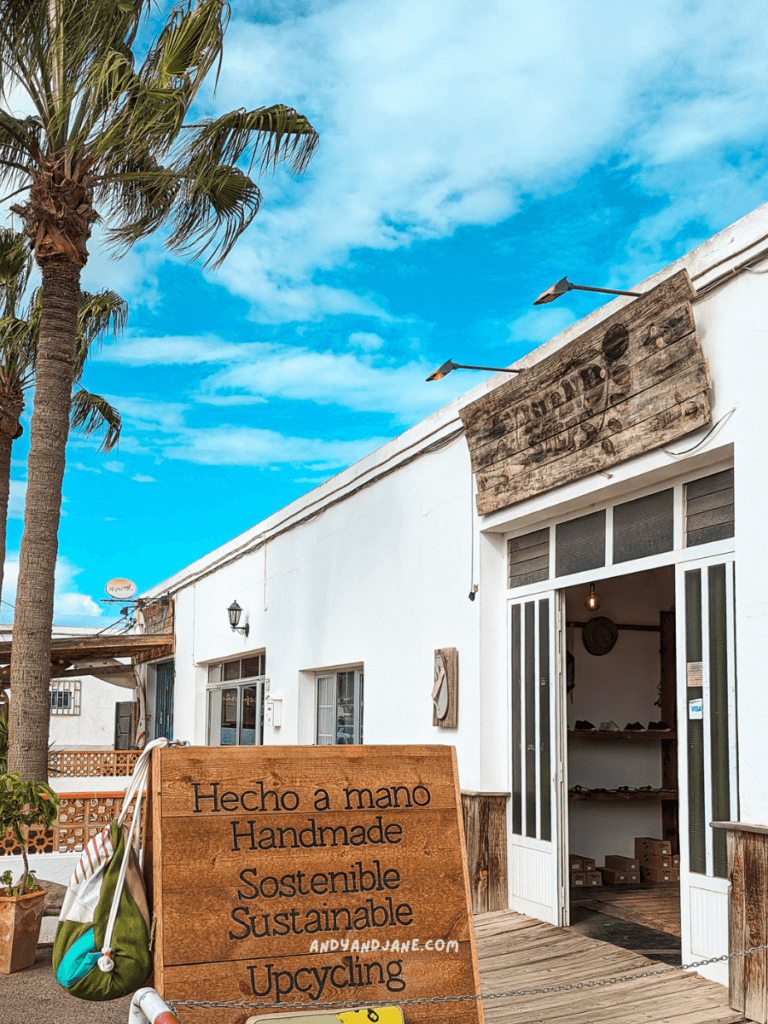 A storefront featuring a wooden sign with "Handmade, Sustainable, Upcycling" in English and Spanish, framed by palm trees and a blue sky in Lajares, Fuerteventura.