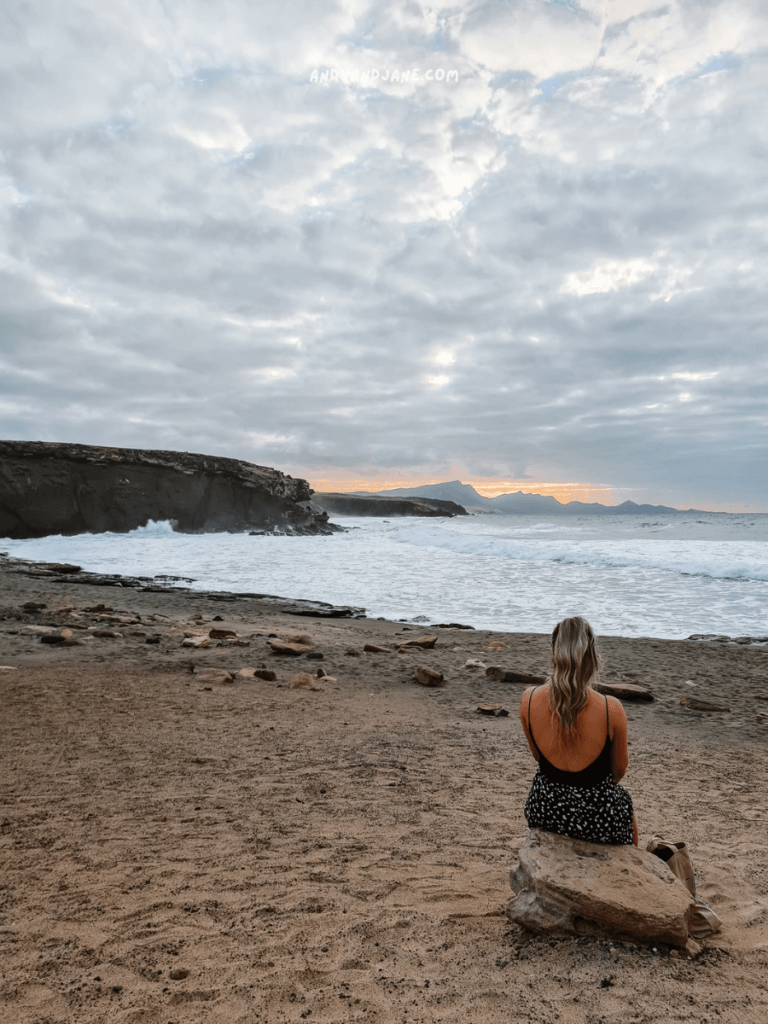 A woman in a black dress sits on a rock by the beach in La Pared, gazing at the ocean waves under a cloudy sky during sunset.