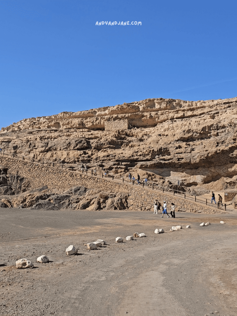 A group of people walk along a stone path leading up a rocky hill under a clear blue sky to the Ajuy Caves in Fuerteventura. White stones line the dusty ground ahead.