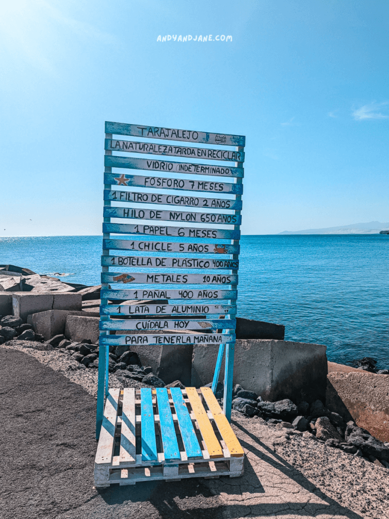 A wooden sign on a beach in Tarajalejo outlines recycling timelines for various materials, highlighting environmental awareness. Sea and sky in background.