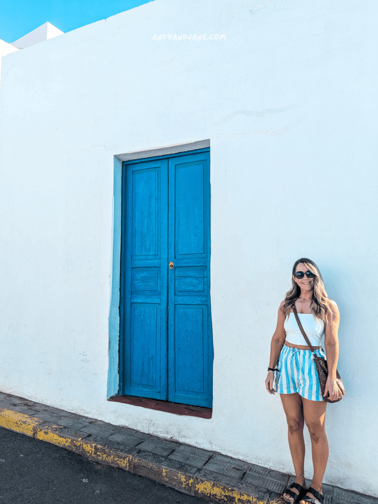 A woman stands beside a vibrant blue wooden door on a whitewashed wall, wearing striped shorts and a tank top, holding a drink.