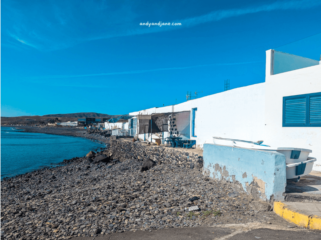 Coastal view of white buildings in Tarajalejo along a rocky shoreline under a clear blue sky, with calm water lapping at the stones.