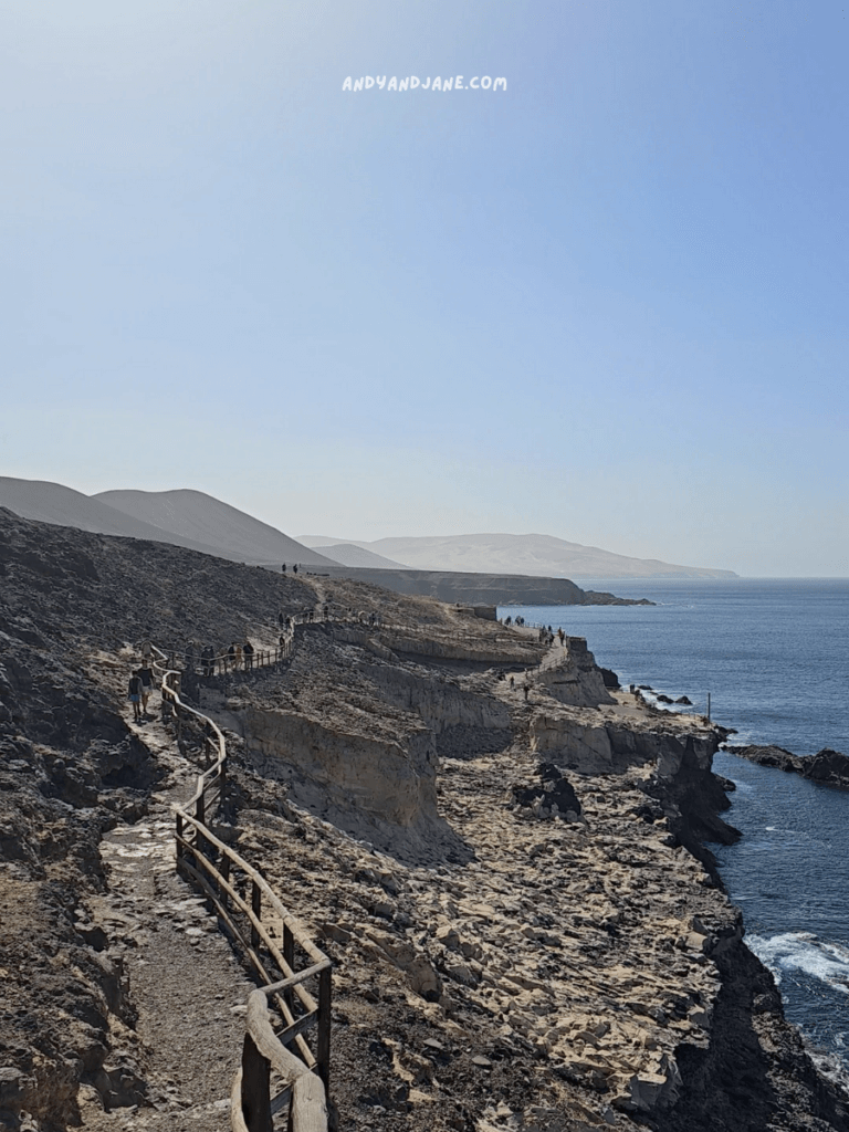 A scenic coastal pathway to the Ajuy Caves in Fuerteventura winding along rocky cliffs, with hikers enjoying the stunning ocean views under a clear blue sky.