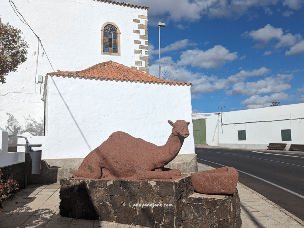 A reddish-brown stone sculpture of a camel, placed on a stone pedestal beside a white building under a blue sky with clouds in Tuineje, Fuerteventura.