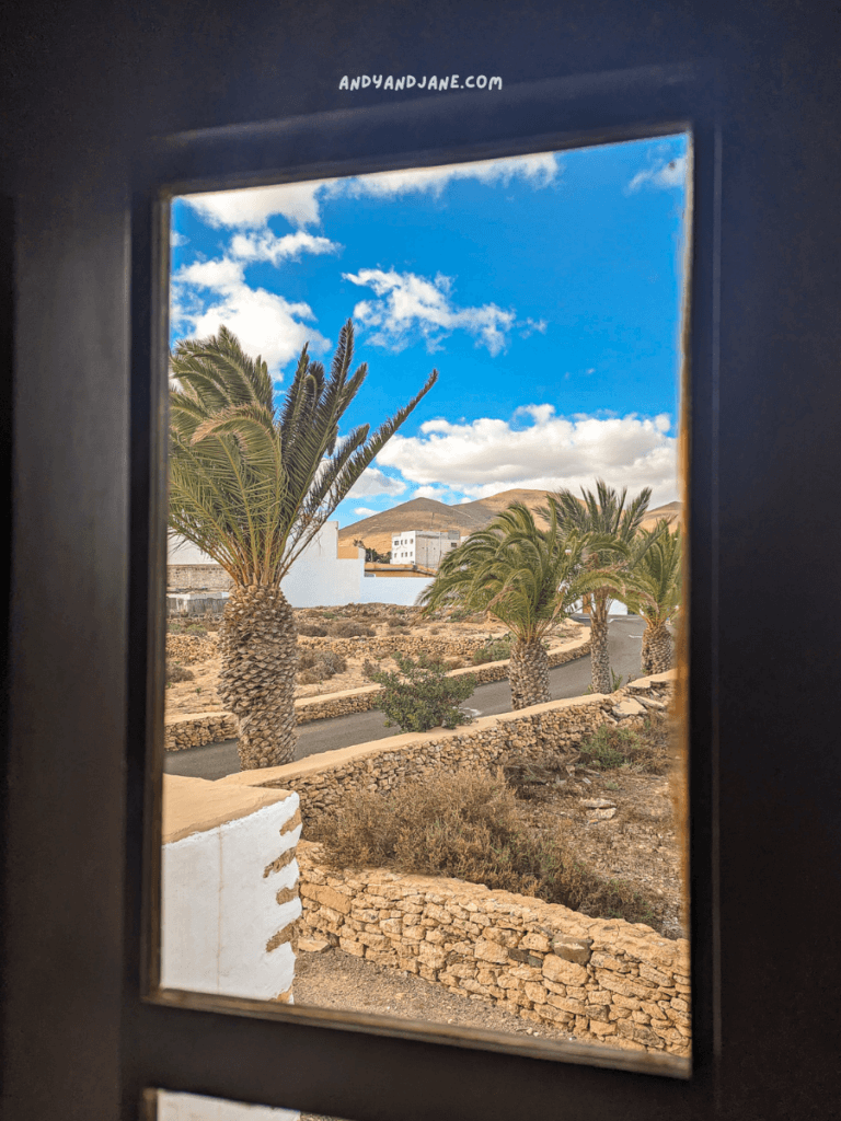 A window view from the museum in Tuineje, showcasing palm trees, a winding pathway, and blue skies with clouds amid a rustic landscape of stone walls.