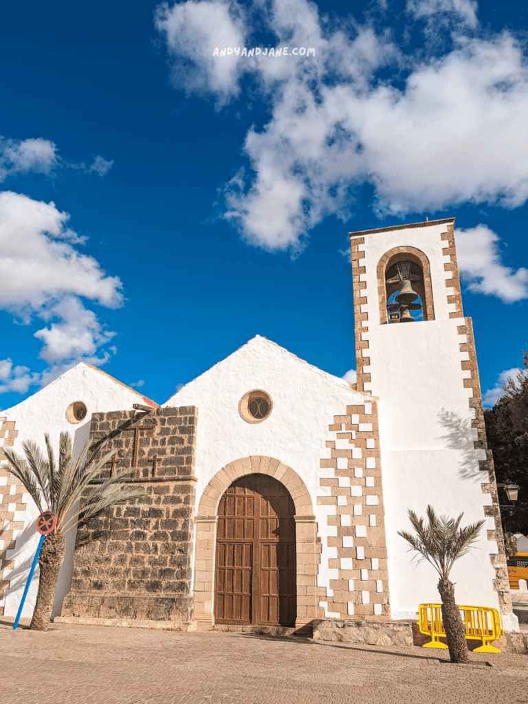 White church with a bell tower in Tuineje, Fuerteventura, surrounded by palm trees, under a blue sky with clouds. 