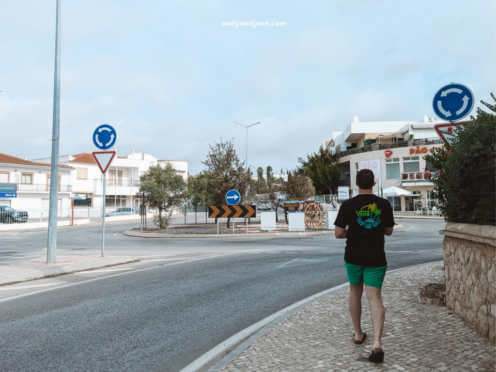 A man wearing a black t-shirt and green shorts walks along a sidewalk near a roundabout in Vale de Parra, Portugal - under a cloudy sky.