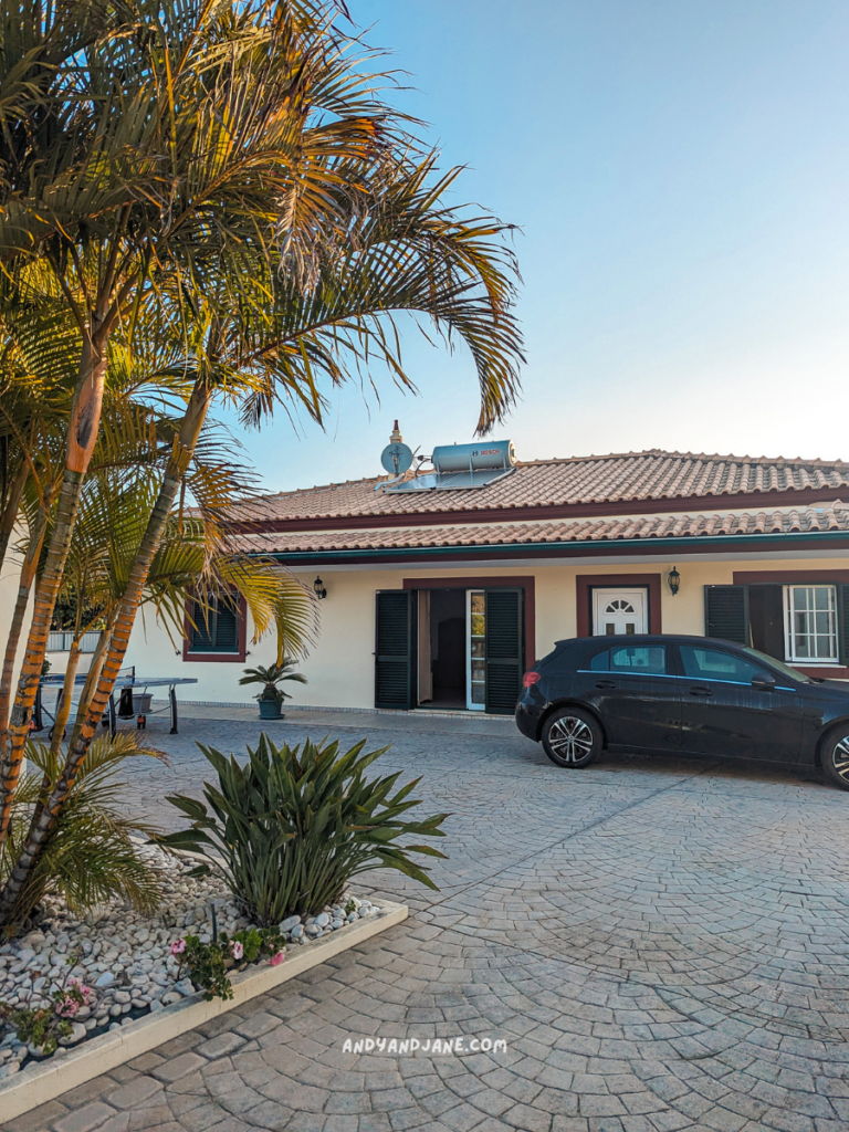 Villa Arvela, Algarve, with a tiled roof, surrounded by palm trees and plants, showing a parked black car in a paved driveway under a clear sky.