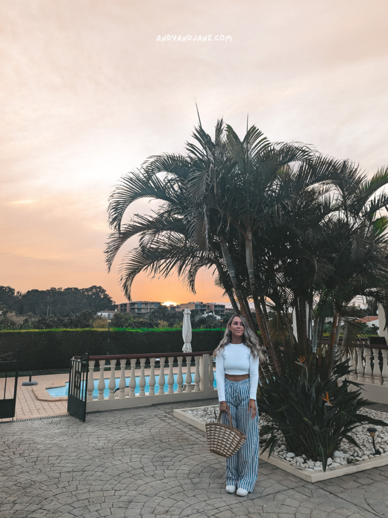 A person stands near a tall palm tree at sunset, wearing a white top and striped pants, holding a woven basket. A pool is visible in the background at Villa Arvela, Gale.