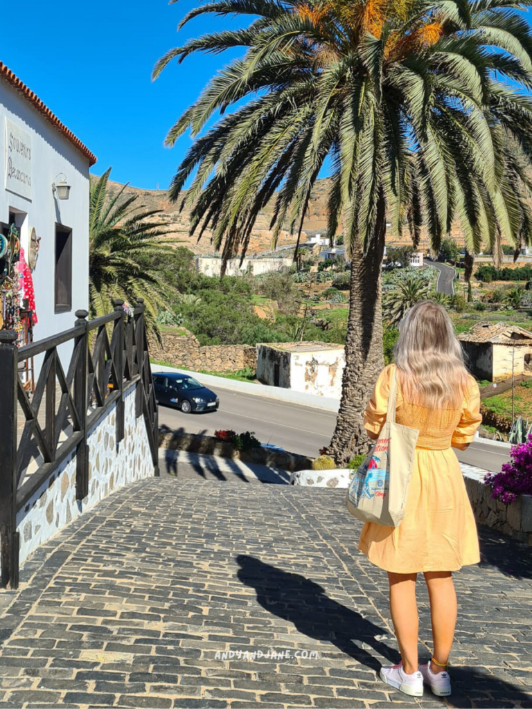 A woman in a yellow dress stands on a cobblestone path, looking towards palm trees and a scenic landscape under a clear blue sky.