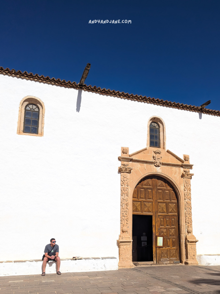 A historic building with a large wooden door and intricate stonework, set against a bright blue sky and sunlit stone pavement.