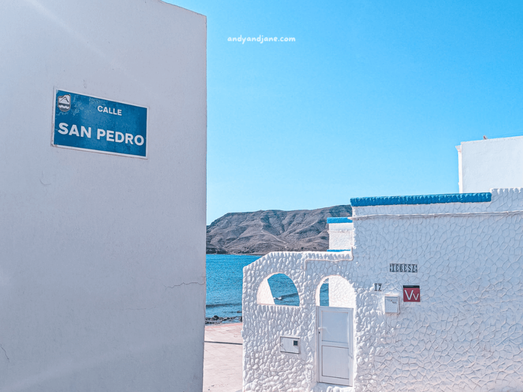 A street sign for "Calle San Pedro" in Las Playitas next to a white, textured building by the ocean, with mountains in the background and clear blue sky.