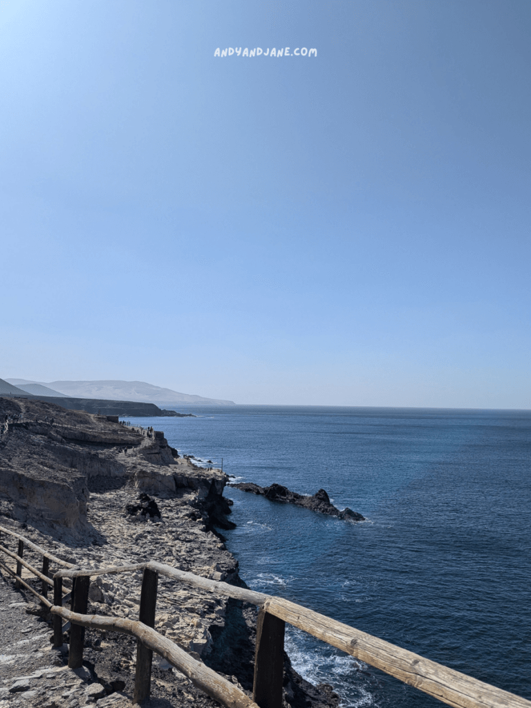 Scenic coastline with rocky cliffs, calm blue waters, and a wooden fence under a clear blue sky, hinting at a tranquil seaside walk.