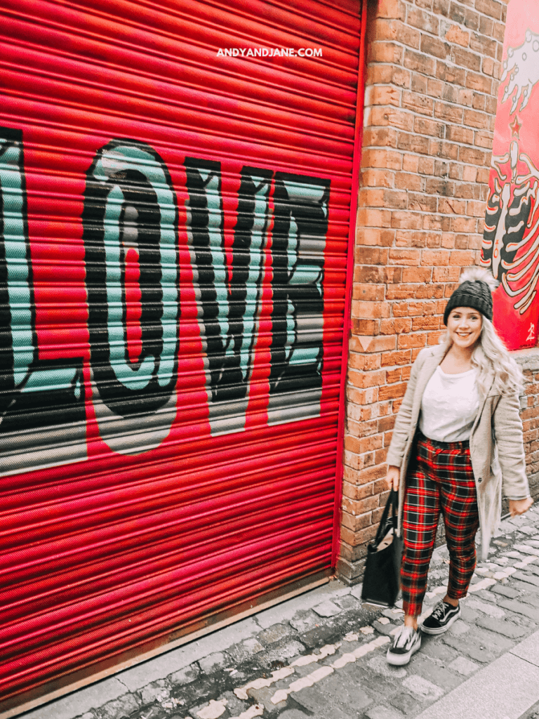 A person in plaid pants walks past a vibrant red roller door with the word 'LOVE' painted in bold black and teal letters in Belfast, Northern Ireland