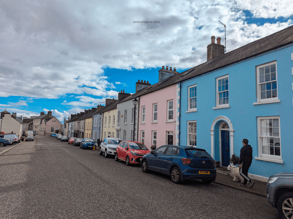 A colorful street lined with pastel houses, parked cars, and a person walking a dog under a partly cloudy sky.