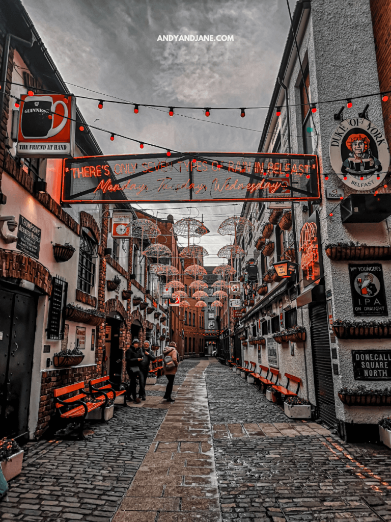 A vibrant Belfast alleyway adorned with umbrellas and string lights, featuring cobblestone paths and colorful pub signs.