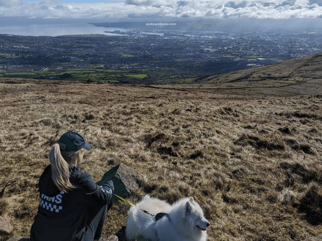 A woman in a black jacket sits on a hilltop, enjoying a scenic view with her fluffy white dog beside her.