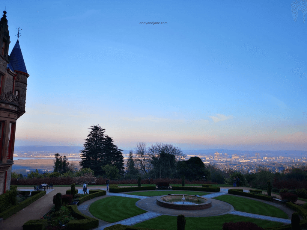 View from a landscaped garden at Belfast Castle featuring a fountain, surrounded by trees, with a historic building and the vast Belfast skyline in the background.