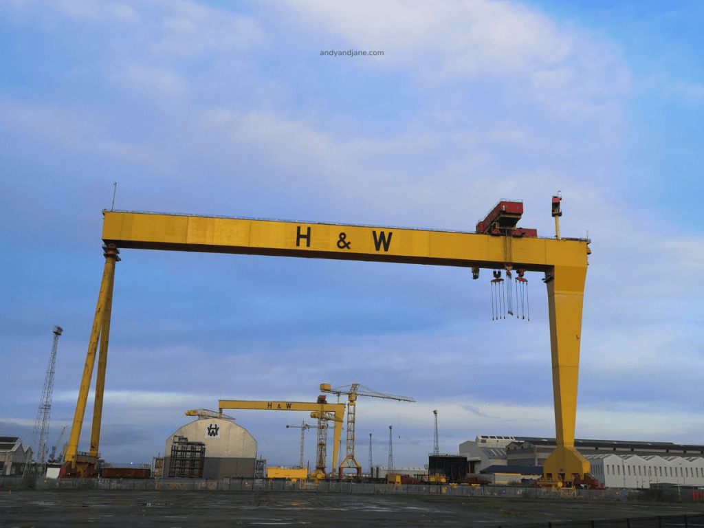 A large yellow gantry crane labelled 'H & W' towers over a shipyard, surrounded by machinery and buildings under a cloudy sky.