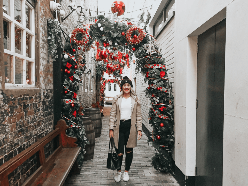 A woman stands in a festive alley in Belfast, adorned with red decorations and greenery, with rustic stone walls and wooden benches.