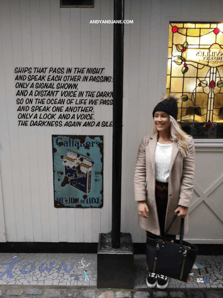 A woman stands in a cozy alley with a vintage sign in Belfast, illuminated by a colorful stained glass window and accompanied by poetic text on the wall.