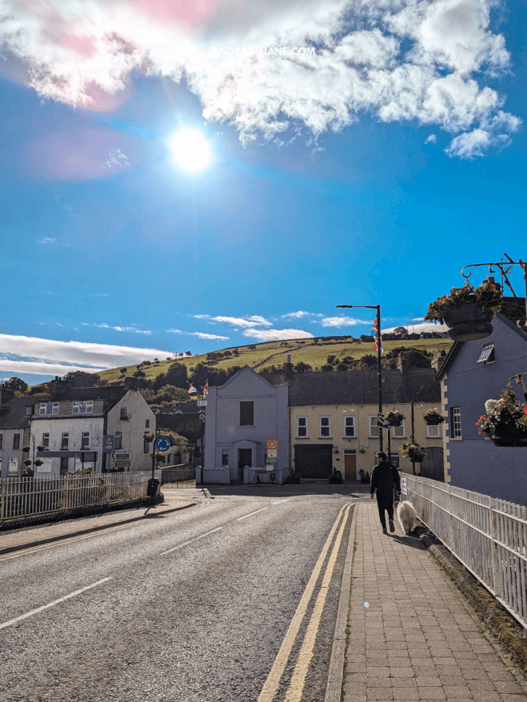 A sunny street scene with a person walking a dog, surrounded by quaint houses and hills under a bright blue sky.