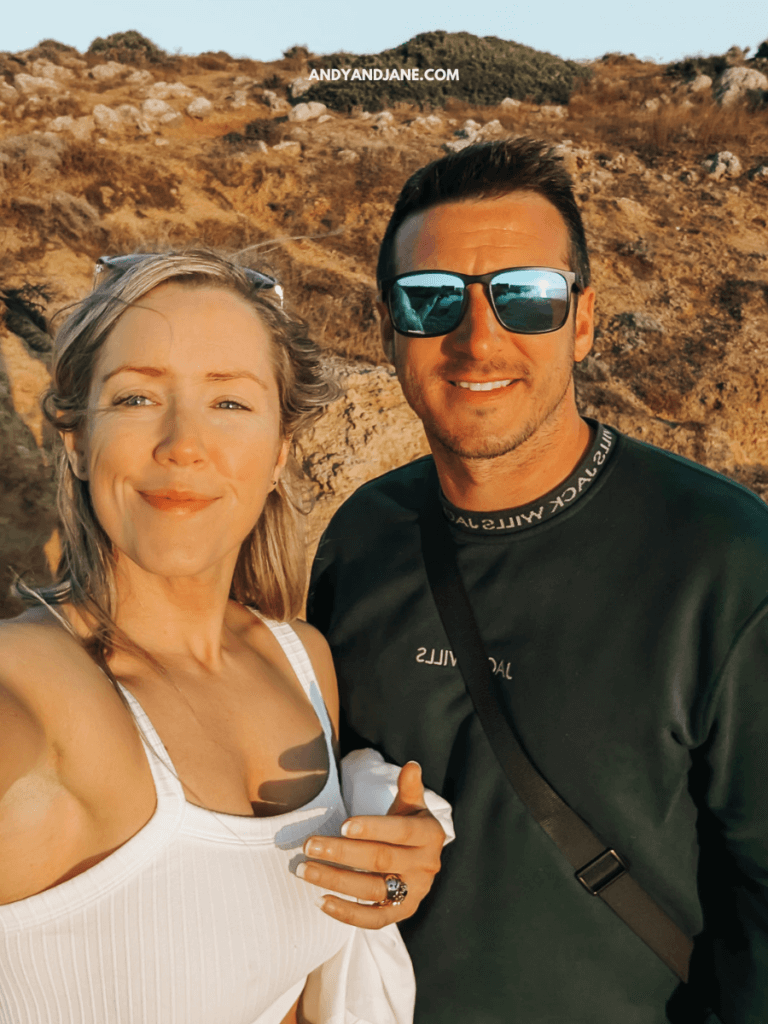 A woman in a white top and a man in a dark sweater pose together in a sunlit outdoor setting with rocky terrain in the background.