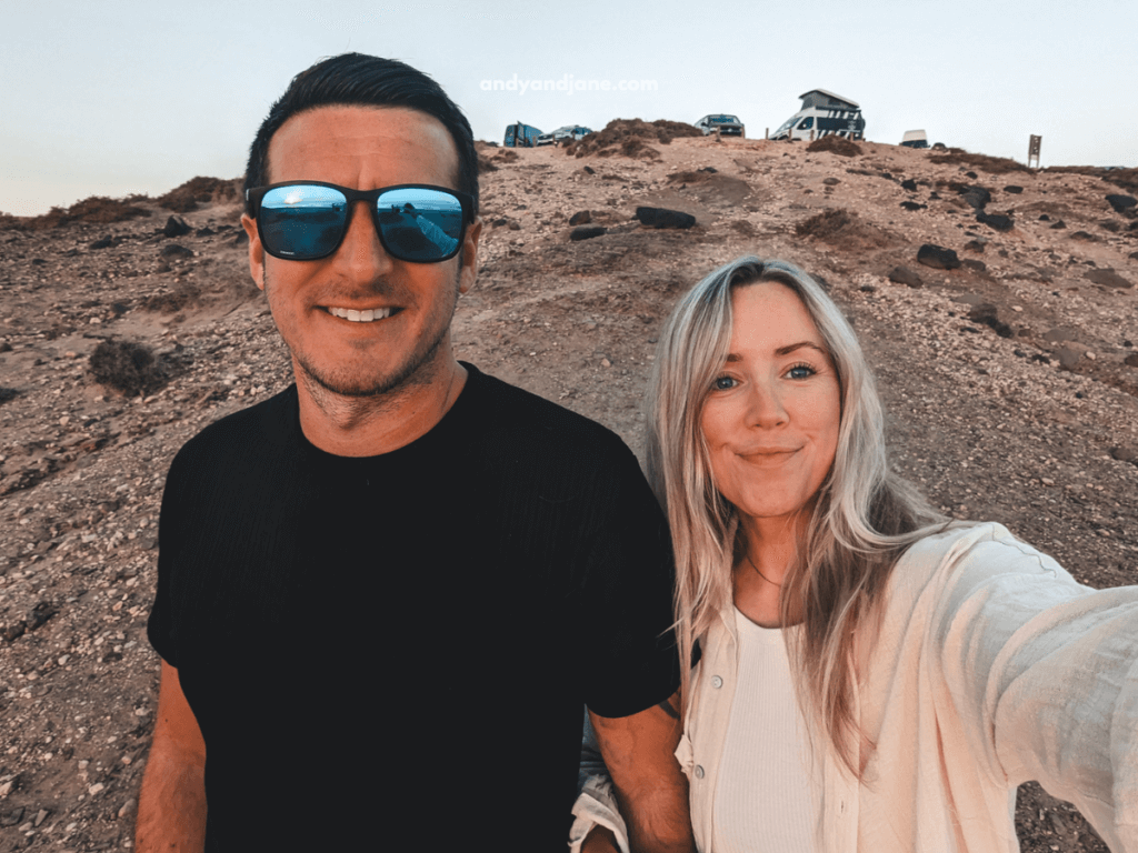 A couple poses for a selfie on a rocky hillside during sunset, surrounded by cars in the background.