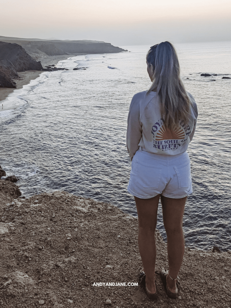 A woman stands on a cliff overlooking a serene ocean at dusk, wearing a surf-themed sweatshirt and white shorts.