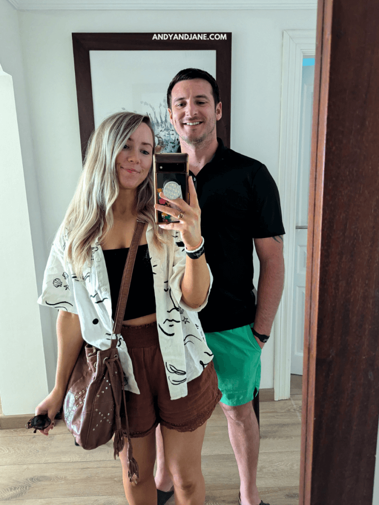 A couple pose for a mirror selfie. The woman wears a patterned shirt and brown shorts, holding sunglasses and a handbag. The man stands beside her.