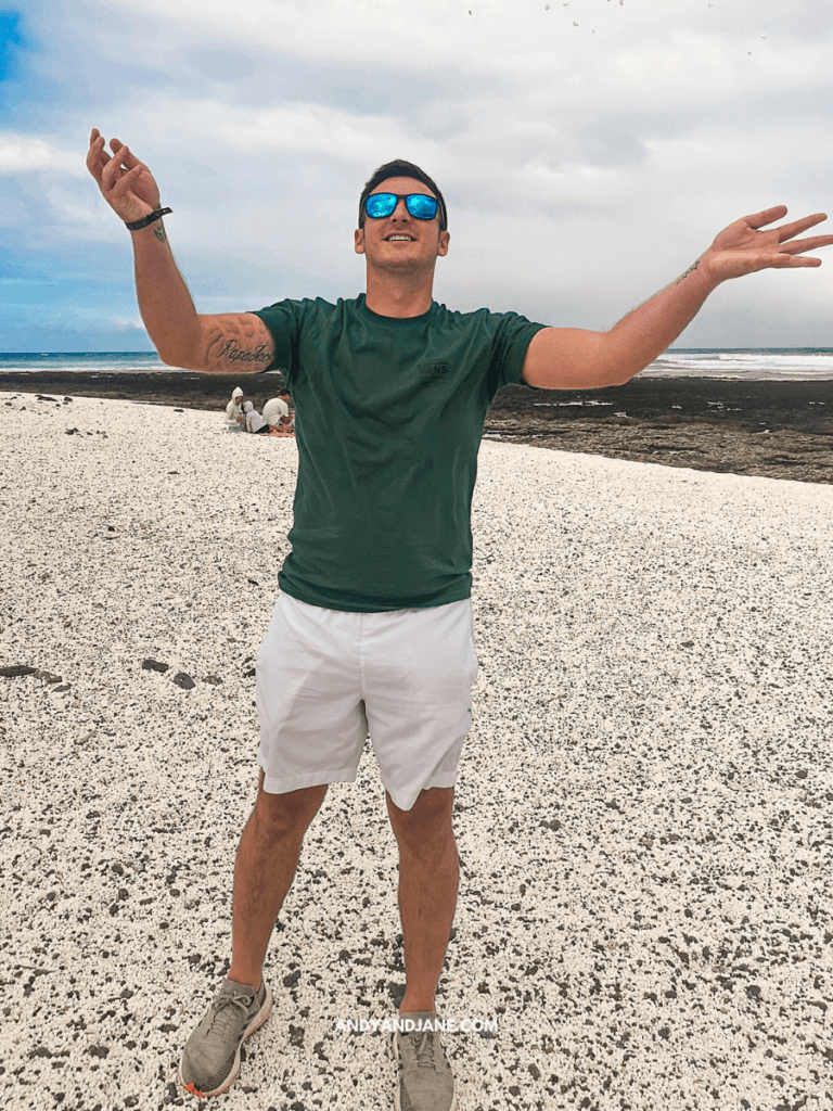 A smiling man in a green shirt and white shorts stands with arms raised on a pebbly beach, with the ocean and cloudy sky in the background.