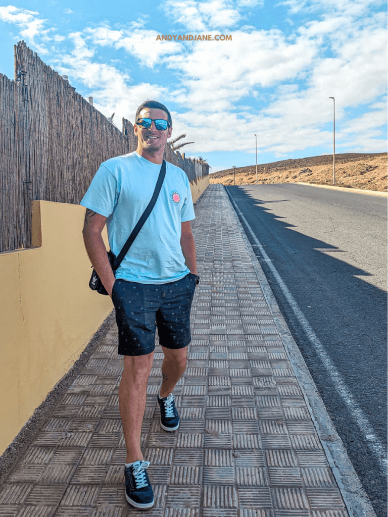 A casually dressed man walks on a tiled sidewalk next to a yellow wall, with a clear blue sky and hilly terrain in the background.