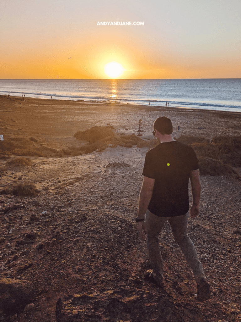 A person walks down a rocky path towards the beach at sunset, with gentle waves and silhouettes of people in the distance.