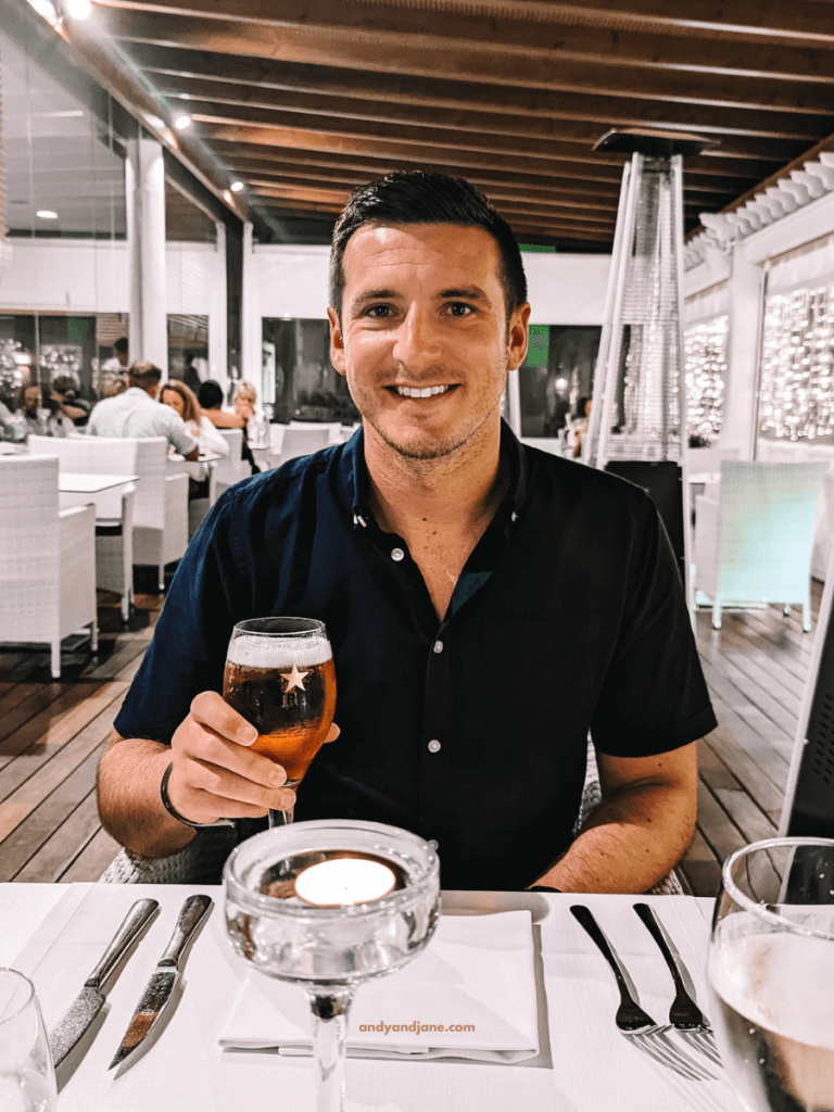 A man holds a beer glass while seated at a dining table, surrounded by elegant decor and soft lighting in a vibrant restaurant atmosphere.