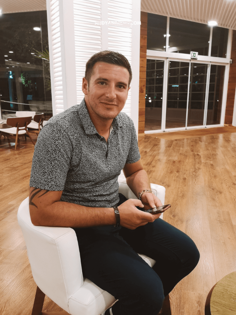 A man sits on a white chair, focused on his phone, in a modern, softly lit setting with wooden floors and bright decor.