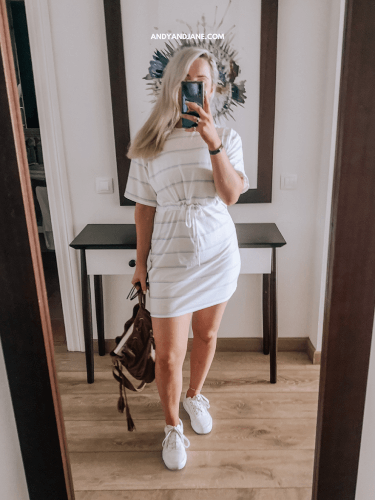 A person wearing a striped dress with a drawstring waist stands in a mirror, holding a brown bag, paired with white sneakers.