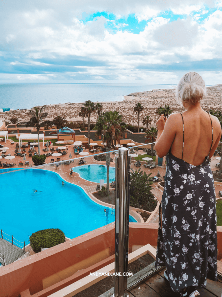 A woman in a floral dress stands on a balcony overlooking a blue pool and the ocean, framed by palm trees under a cloudy sky.