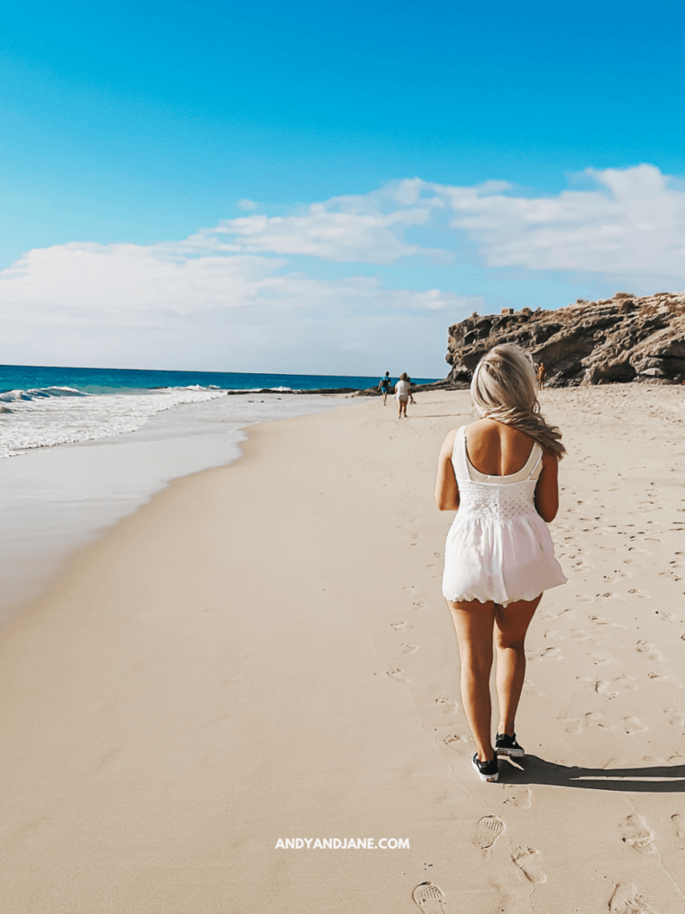 A woman in a white dress walks along a sandy beach, with the ocean waves and rocky cliffs in the background under a blue sky.