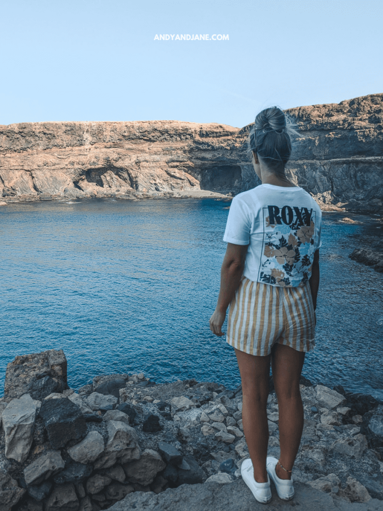 A woman in a Roxy shirt and striped shorts stands on rocky cliffs, gazing at a tranquil blue sea and distant cliffs under a clear sky.