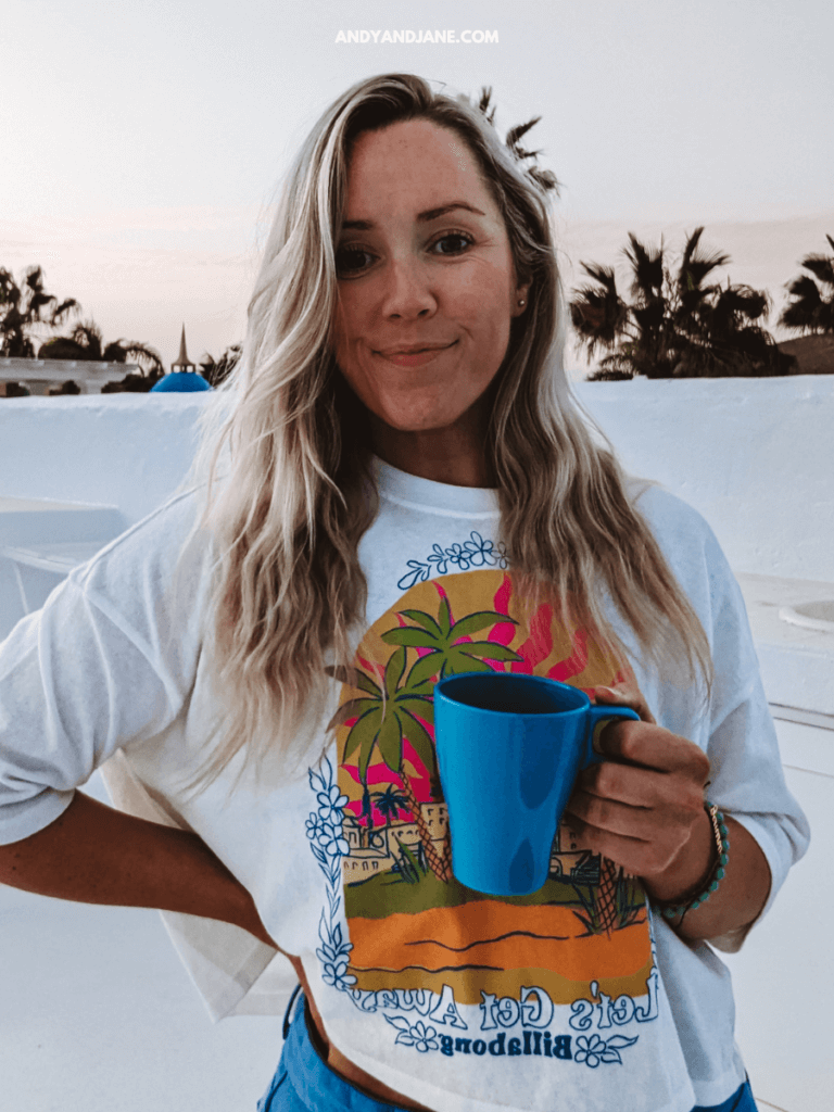 A person stands outdoors holding a blue mug, wearing a colorful graphic t-shirt, with palm trees and a sunset in the background.