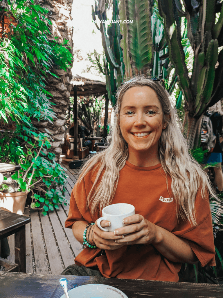 A person with long, wavy hair sits at a wooden table surrounded by tropical plants, holding a white coffee cup.