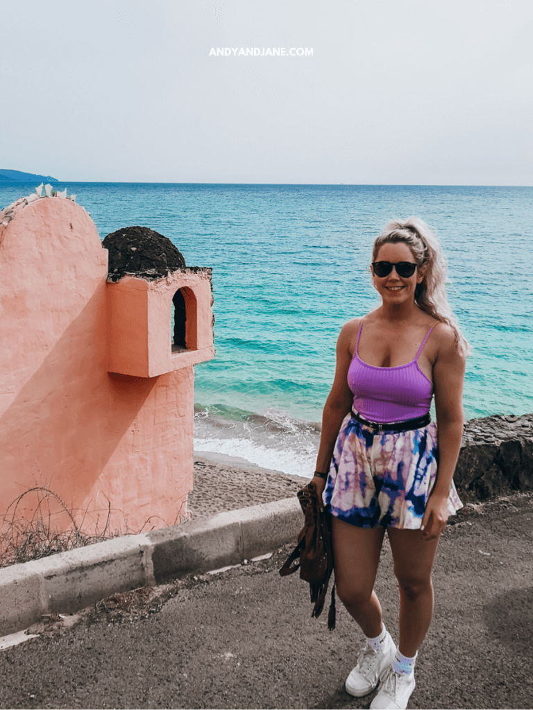 A woman stands on a coastal road near a pink building, with turquoise waves lapping against a sandy beach in the background.