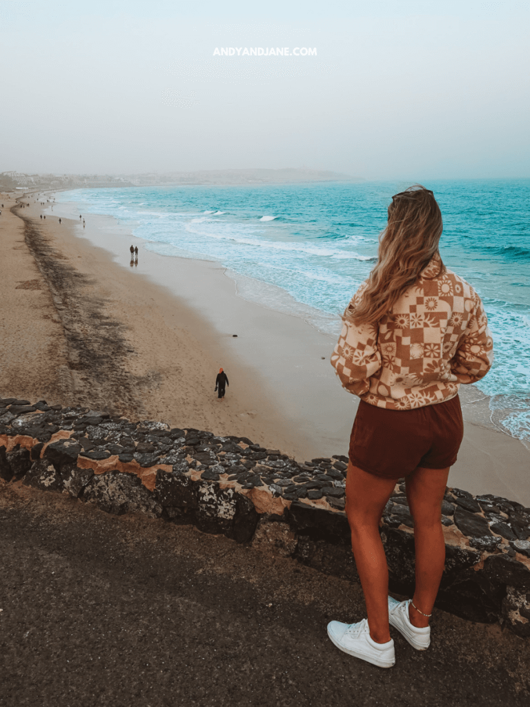 A woman in a patterned sweater and shorts gazes out at a beach with gentle waves and people walking along the shore.